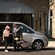 A woman wearing a visor stands by a van with tinted windows and a man in a suit carrying a bag of golf